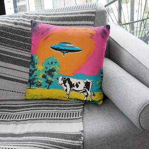 May include: Square throw pillow with a pop art design featuring a black and white cow in a field, a blue UFO, and a colorful sunset. The pillow is placed on a gray sofa.