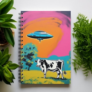 Puede incluir: Una libreta de espiral con una cubierta colorida que presenta una vaca en un campo, un platillo volador en el cielo y una puesta de sol rosa, naranja y azul.