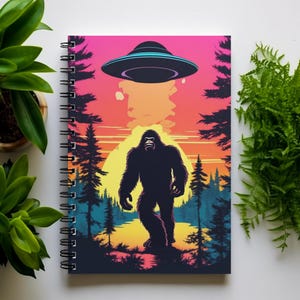 May include: A spiral bound notebook with a colorful illustration of Bigfoot standing in a forest with a UFO flying overhead. The background is a sunset with a pink, orange, and yellow color scheme.