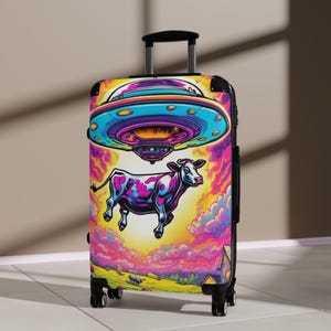 May include: A colorful suitcase with a psychedelic design featuring a cow being lifted by a UFO. The suitcase has a black handle and wheels.