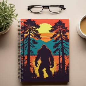 May include: A spiral bound notebook with a colorful illustration of Bigfoot standing in a forest at sunset. The illustration features a silhouette of Bigfoot against a backdrop of trees, mountains, and a lake. The colors are vibrant and include shades of orange, yellow, blue, and green.