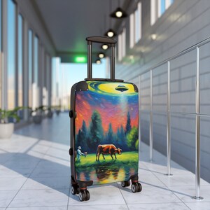 May include: A rolling suitcase with a colorful painting of a cow in a field with a UFO above. The artwork features a vibrant sunset, trees, and a figure in a blue suit. The suitcase has a black handle and wheels.