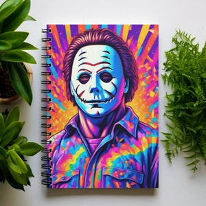 May include: A spiral bound notebook with a colorful illustration of a man wearing a white mask with a scary smile. The background is a vibrant mix of pink, orange, yellow, blue, and purple.