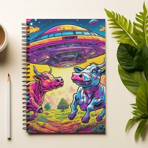 Puede incluir: Una libreta de espiral con una ilustración colorida de dos vacas y un platillo volante. Las vacas son rosadas y azules con cuernos y pezuñas negras. El platillo volante es amarillo con la parte inferior morada. El fondo es un paisaje psicodélico con estrellas y nubes.