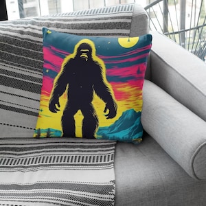 Può includere: Un cuscino con uno sfondo di tramonto colorato e una silhouette di Bigfoot in piedi di fronte a una luna piena. Il cuscino è su un divano grigio con una coperta a righe bianche e nere.