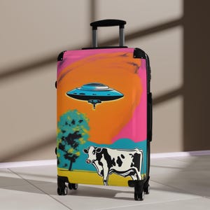 May include: A pink, orange, blue, and green suitcase with a cartoon cow and a flying saucer on it.