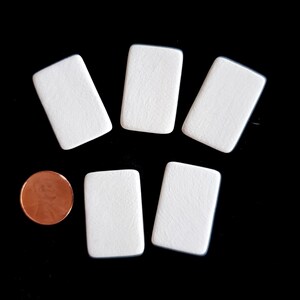 May include: Five small, rectangular, white wooden pieces with rounded corners arranged in a group. A single copper-colored United States one-cent coin is positioned to the left of the bottom row for scale, showing the relative size of the items.