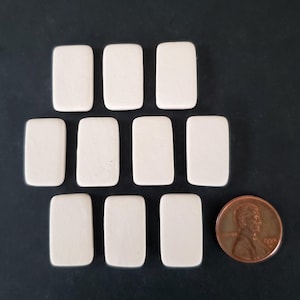 May include: A set of nine small, rectangular, off-white tiles with rounded corners, arranged in three rows. A copper-colored United States one-cent coin is positioned to the right of the bottom row for scale, measuring 0.75 inches in diameter.