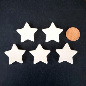 May include: Five smooth, off-white, five-pointed star-shaped objects arranged in a group. A single copper-colored circular coin is positioned to the right of the top right star, providing a visual reference for the size of the stars.