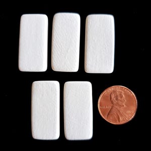May include: Five rectangular white wooden pieces with rounded corners arranged in a group next to a copper-colored United States one-cent coin for scale. The pieces have a flat, matte surface and a uniform, solid white color throughout.