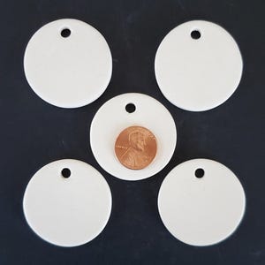 May include: Five white circular ceramic tags, each featuring a single hole at the top for hanging. A copper-colored United States one-cent coin is placed on the center tag to provide a visual scale reference for the size of the circular discs.