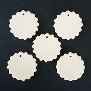 May include: Five off-white ceramic ornaments, each shaped like a circle with a scalloped edge. Every ornament features a small, centered hole near the top rim, designed for hanging. The surfaces are smooth, matte, and uniform in color.