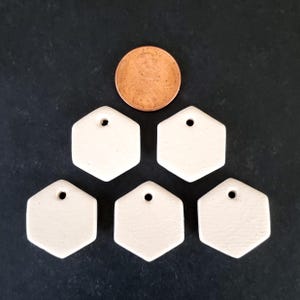 May include: Five small, off-white, matte-finish ceramic hexagon charms, each featuring a single pre-drilled hole at the top. A copper-colored United States penny is positioned above the charms to provide a clear sense of scale.