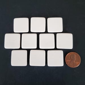 May include: A collection of nine small, smooth, off-white square tiles arranged in three rows. A single copper-colored United States one-cent coin is positioned to the right of the bottom row to provide a visual scale for the tiles.