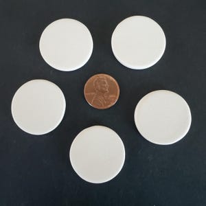 May include: Five plain white circular discs arranged in a pentagonal pattern around a single copper-colored United States one-cent coin placed in the center. Each disc has a smooth, matte surface and a consistent, flat, round shape.