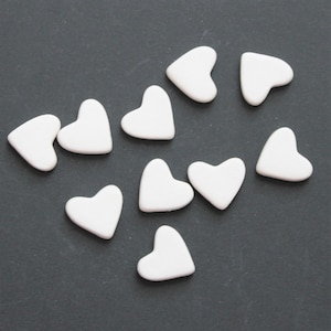 May include: Ten white ceramic hearts on a dark gray background. The hearts are all different sizes and shapes.