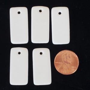 May include: Six white rectangular ceramic tags with a hole at the top, arranged on a black surface. A copper penny is next to the tags for size comparison. The tags have rounded corners and a slightly textured surface.
