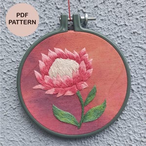 Puede incluir: Un bastidor de bordado circular que contiene una pieza de tela con una flor de protea rosa y blanca bordada a mano y hojas verdes. La tela de fondo presenta un degradado de rojo a naranja. El texto PDF PATTERN es visible arriba a la izquierda.