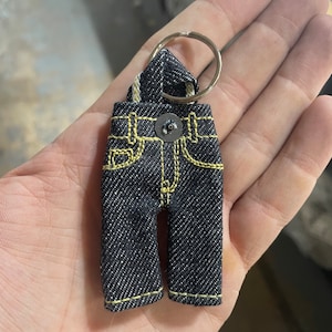 Może przedstawiać: Mały brelok z dżinsu w kształcie kombinezonu z żółtymi szwami i srebrnym guzikiem.