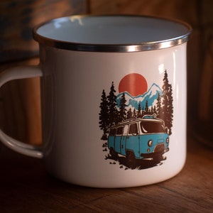 Puede incluir: Taza de camping de esmalte blanco con un gráfico de furgoneta vintage azul, montañas, árboles y un sol rojo. La taza tiene un borde plateado.