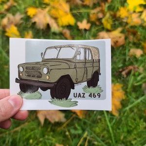 May include: A sticker featuring a cartoon illustration of a UAZ 469 military vehicle in olive green, with black tires and accents. The sticker is held against a backdrop of green grass and fallen leaves.