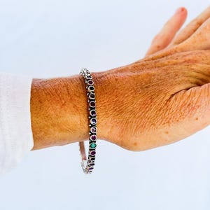 May include: A silver-toned bangle bracelet featuring a continuous row of small, round, bezel-set gemstones in alternating shades of deep red, dark purple, and a single vibrant green stone, worn around the wrist against a white background.