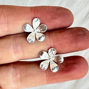 Puede incluir: Dos pequeños pendientes de botón plateados con forma de flor. Cada pendiente tiene cinco pétalos y una superficie texturizada. Los pendientes se muestran en los dedos de una persona sobre un fondo blanco.