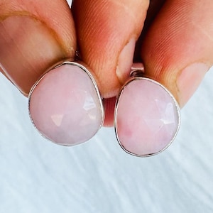 May include: A pair of stud earrings featuring faceted, teardrop-shaped light pink stones set in polished silver-toned metal frames. The stones have a smooth, reflective surface with multiple geometric facets that catch the light.