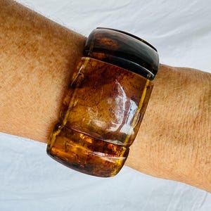 May include: A wide, polished cuff bracelet featuring a translucent, deep amber-colored resin with swirling dark brown and golden inclusions. The bracelet has a smooth, rounded surface and a thick, substantial profile that wraps around the wrist.