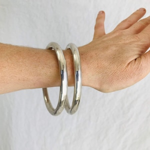 May include: Two silver bangle bracelets stacked on a person's wrist. The bracelets are smooth and shiny.