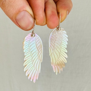 May include: Iridescent wing-shaped earrings with silver hooks. The earrings have a layered feather design and reflect a rainbow of colors. These unique earrings are a beautiful accessory.