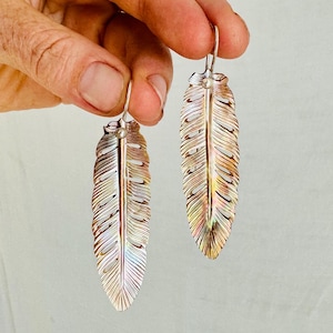 Op de afbeelding: Een paar zilverkleurige oorringen in de vorm van veren. Elke oorring heeft een lange, slanke veervorm met gedetailleerde adering. De veren hebben een kleurengradiënt van zilver naar goud. De oorbellen zijn opgehangen aan een kleine haak.