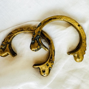 May include: Two antique brass bracelets with ornate, curved designs. The bracelets have a weathered, aged appearance, with dark spots and a golden hue. They are resting on a white, textured fabric.