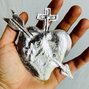 May include: A silver-colored heart-shaped religious pendant pierced by an arrow, with a cross at the top. The pendant is held in a hand, showcasing its detailed design and metallic sheen. The heart is approximately 3 inches wide.