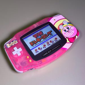 May include: A translucent pink Game Boy Advance handheld gaming console featuring a Kirby character graphic on the right side and the text Star Kirby in Japanese on the left. The screen displays the Mario vs. Donkey Kong game title screen.