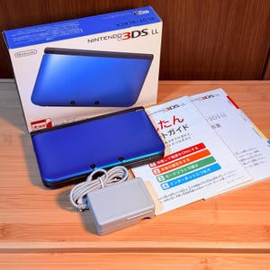 May include: A blue and black Nintendo 3DS LL handheld gaming console with its original white packaging box, a grey power adapter, and Japanese instruction manuals. The console features a metallic blue top cover and a black base, shown closed.