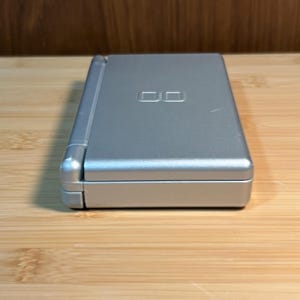 May include: A silver handheld gaming console in a closed, rectangular clamshell design. The top surface features a debossed DS logo centered on the lid. The device has a smooth, metallic finish with a hinge running along the left side of the unit.