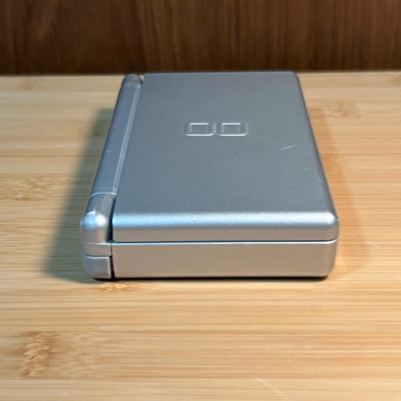 May include: A silver handheld gaming console in a closed, rectangular clamshell design. The top surface features a debossed DS logo centered on the lid. The device has a smooth, metallic finish with a hinge running along the left side of the unit.