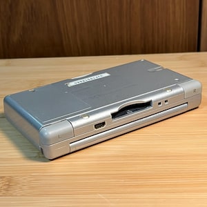 May include: A silver handheld gaming console featuring an L and R shoulder button, a central game cartridge slot, and a charging port. A white rectangular sticker with the serial number UJF19519664 is affixed to the top surface of the device.