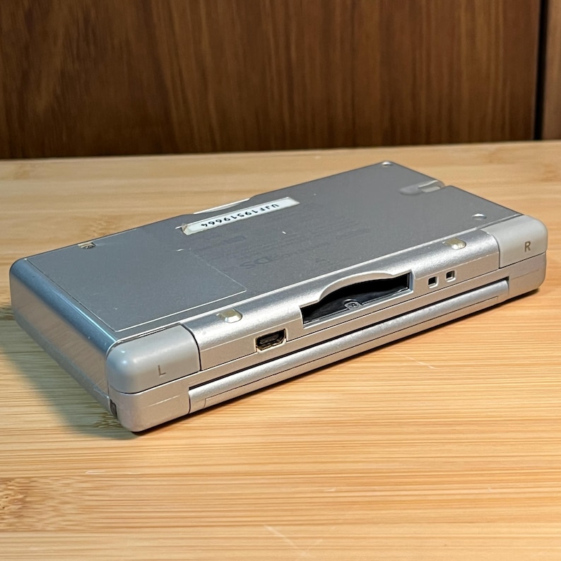 May include: A silver handheld gaming console featuring an L and R shoulder button, a central game cartridge slot, and a charging port. A white rectangular sticker with the serial number UJF19519664 is affixed to the top surface of the device.