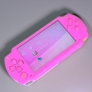 May include: A bright pink handheld gaming console with a rectangular screen displaying a digital menu interface. The device features a directional pad, four action buttons, a joystick, and various control buttons labelled Sony and PSP.