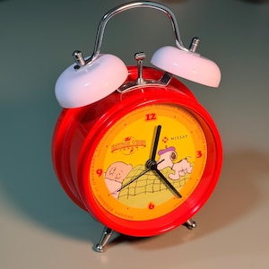 May include: A bright red twin-bell alarm clock featuring a yellow clock face with Snoopy and Woodstock illustrations. The face includes the Universal Studios Japan logo, the Nissay logo, and black clock hands. It has white bells and silver legs.