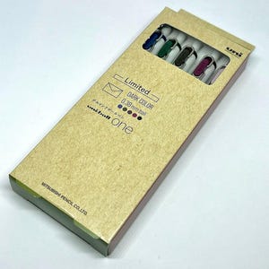 May include: A rectangular cardboard box containing Uni-ball One gel ink pens. The box is tan and features the text "Limited" and "DARK COLOR 0.38 mm ball." The pens are visible through a window and have coloured ink tips.