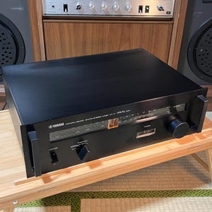 May include: A black Yamaha Natural Sound AM/FM stereo tuner. The front panel features a tuning dial, function selector, and power button. The tuner has a silver and orange display and is sitting on a wooden table.