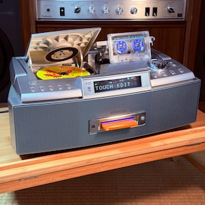 Könnte beinhalten: Ein silbernes Kenwood-Stereosystem mit einem Top-Loading-CD-Player mit gelber Disc, einer oben eingelegten Kassetten-Reinigungskassette und einem Frontschlitz mit einer orangefarbenen Karte. Das Digitaldisplay zeigt den Text TOUCH EDIT.