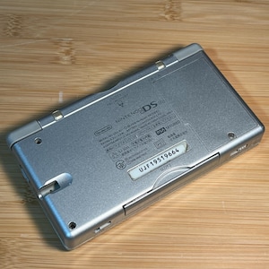 May include: A silver Nintendo DS Lite handheld game console viewed from the back. The casing features printed technical specifications, regulatory markings, and a white label with the serial number UJF19519664. It includes slots labeled SLOT-1 and SLOT-2.