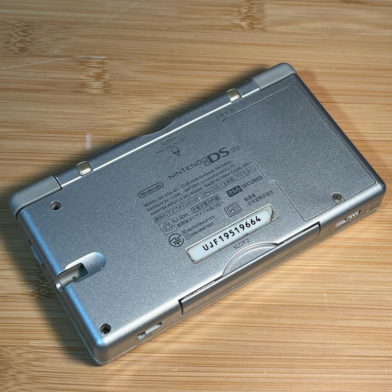 May include: A silver Nintendo DS Lite handheld game console viewed from the back. The casing features printed technical specifications, regulatory markings, and a white label with the serial number UJF19519664. It includes slots labeled SLOT-1 and SLOT-2.