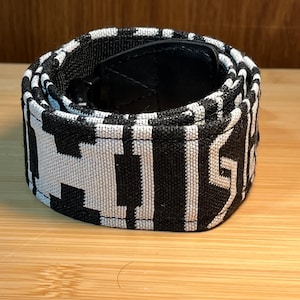 May include: A coiled fabric belt featuring a high-contrast black and white geometric pattern. The strap is constructed from a woven textile material and includes a black buckle mechanism visible at the center of the rolled-up accessory.