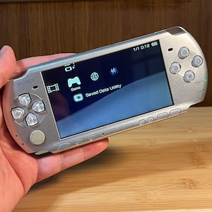 May include: Silver Sony PSP handheld game console. The screen displays a blue and white gradient with icons for Game and Saved Data Utility. The console has a directional pad, buttons, and a power switch. The screen displays the time as 0:12.