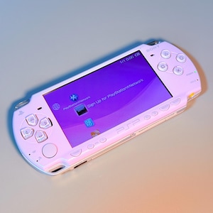 Könnte beinhalten: Eine tragbare Sony PSP-Spielkonsole in hellrosa Ausführung. Das Gerät verfügt über einen Breitbildschirm, der die PlayStation Network-Oberfläche mit blauen Symbolen und Text anzeigt, sowie standardmäßige Richtungstasten und Symbole.